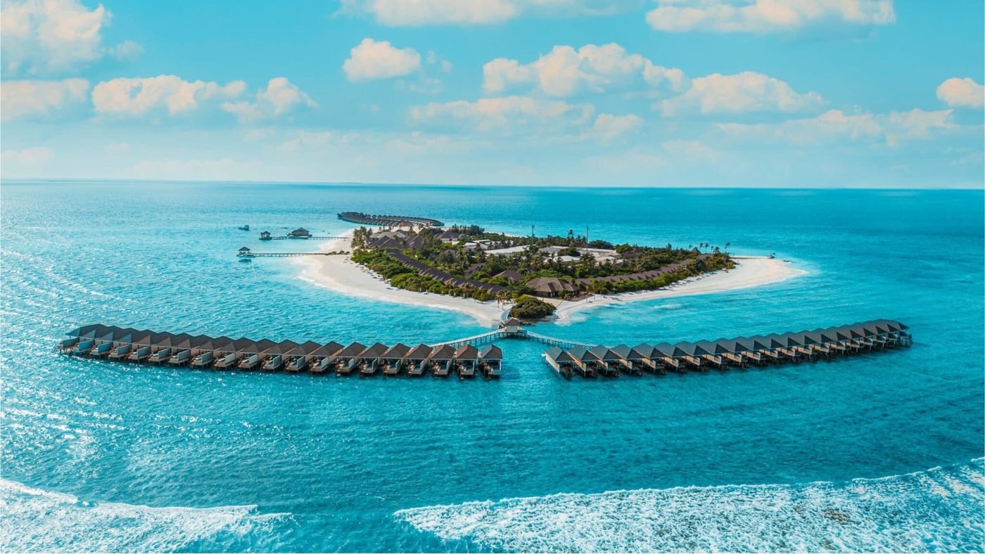 Aerial view of a Maldives island resort with overwater villas and a blue sky